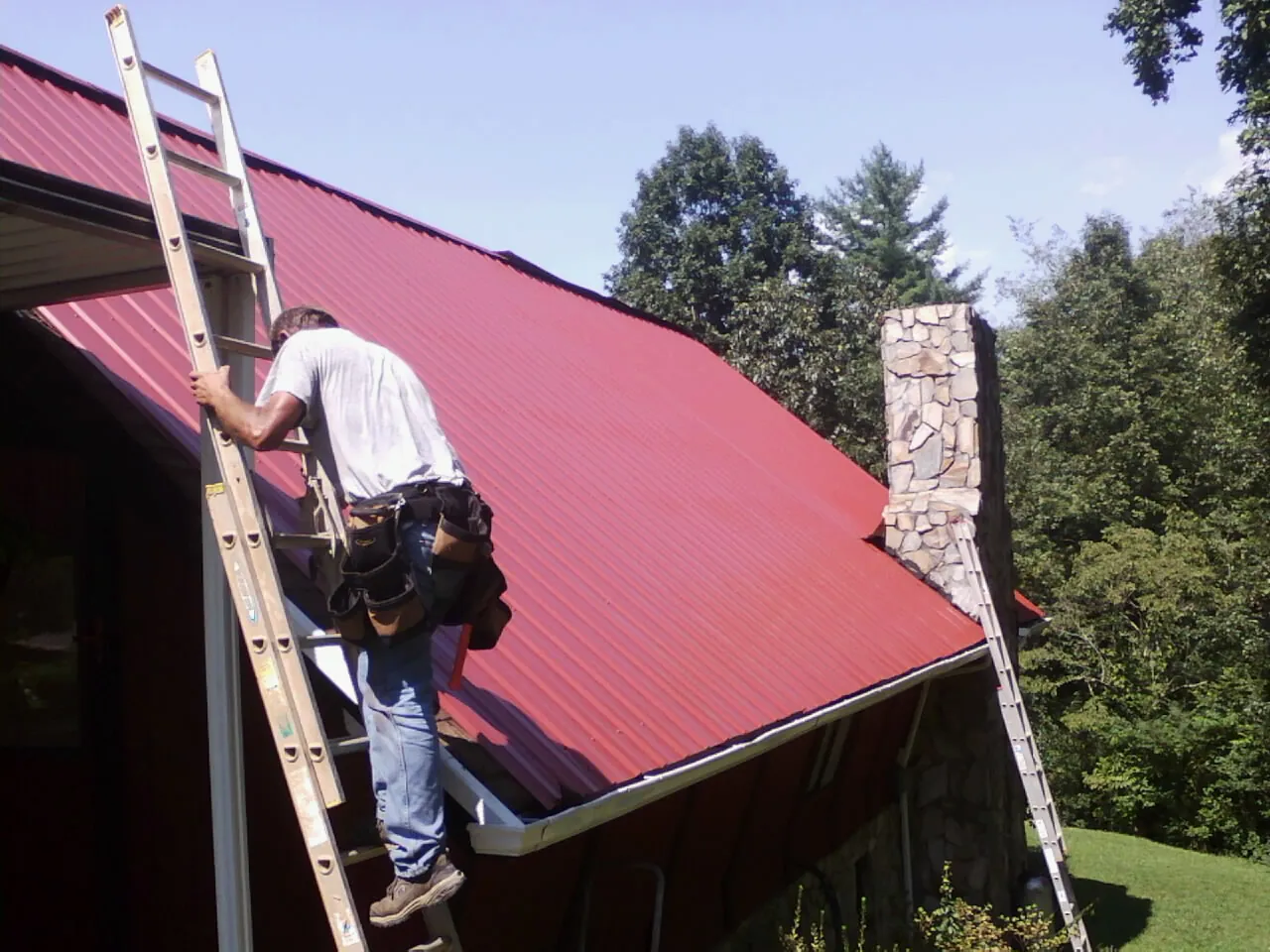 Roofer on a ladder beside a red metal roof and stone chimney — Soffit & Fascia Repair by Roofing Brothers in Albrightsville